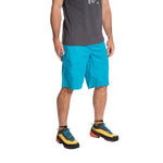 A man stands against a white background wearing La Sportiva Bleauser Shorts Men's in bright blue, featuring a gusseted crotch for comfort. He also wears a dark grey organic cotton t-shirt and colorful hiking shoes. Image is cropped above the shoulders.