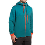 A man wears the La Sportiva Method Hoody Men's—a teal, textured jacket made from recycled fabrics with orange zipper, drawstrings, and accents, subtle chest logo, and modern casual style—paired with black pants.