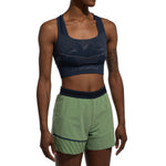 A person models the La Sportiva Focus II Top Women's in dark blue with body-mapped support and geometric patterns, paired with light green shorts featuring dark blue trim, standing against a white background. Their head is out of frame.