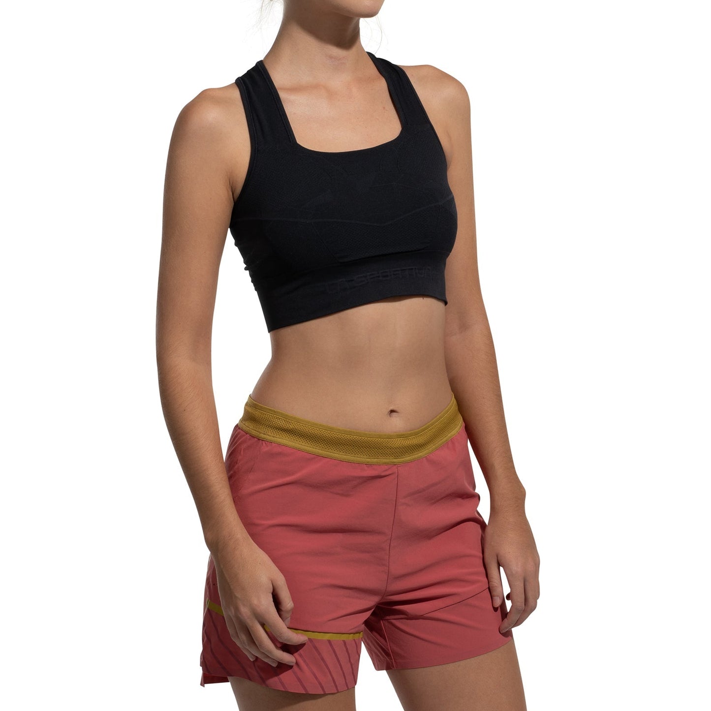 A person wears the La Sportiva Focus II Top Women's, a black sports bra with body-mapped support, paired with pink athletic shorts with a yellow waistband. They stand against a white background, face not visible.