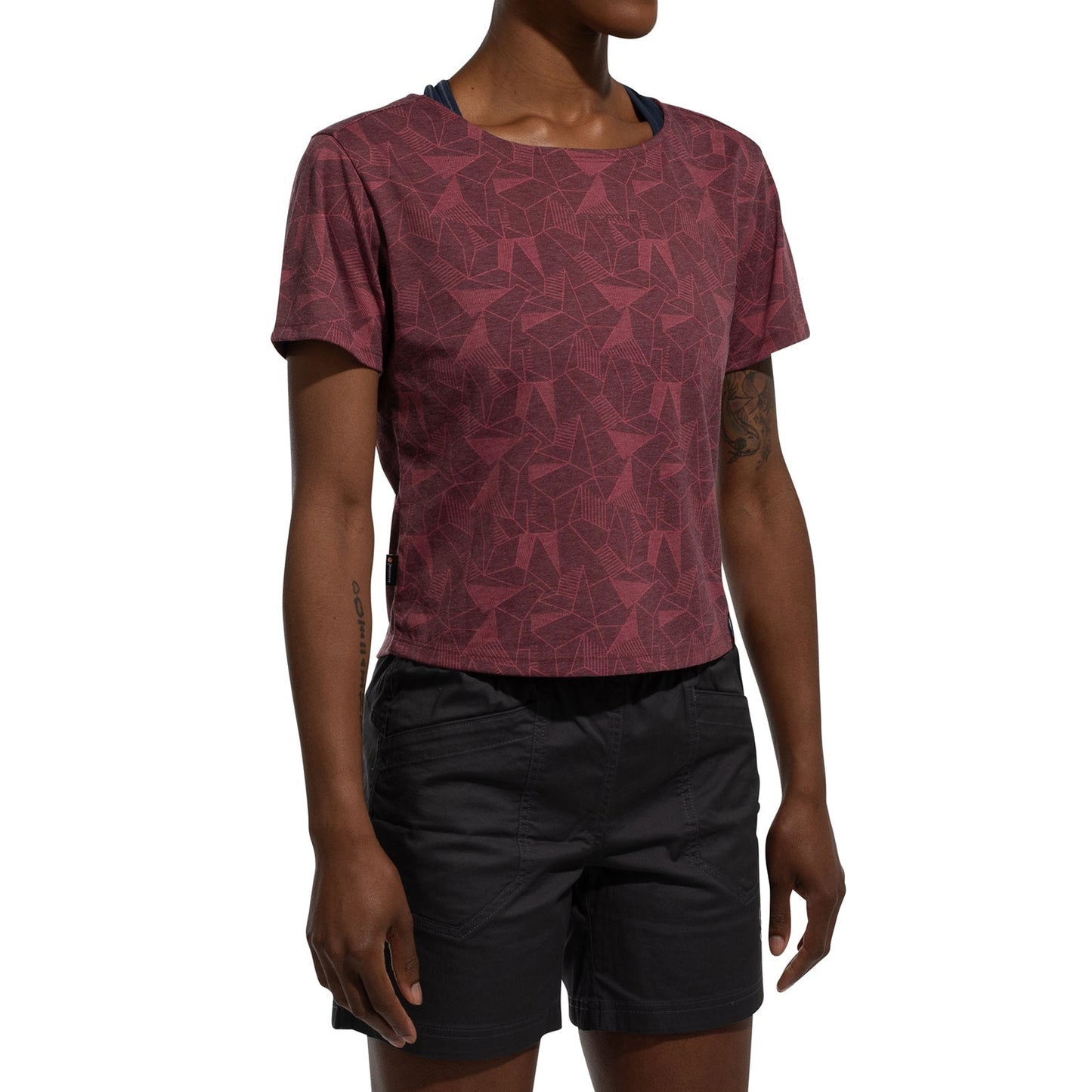 A person stands against a white background, arms relaxed, wearing the La Sportiva Dimension T-Shirt Women's (2025) in maroon with a pattern and black baggy shorts. Their head is slightly turned and not fully visible.