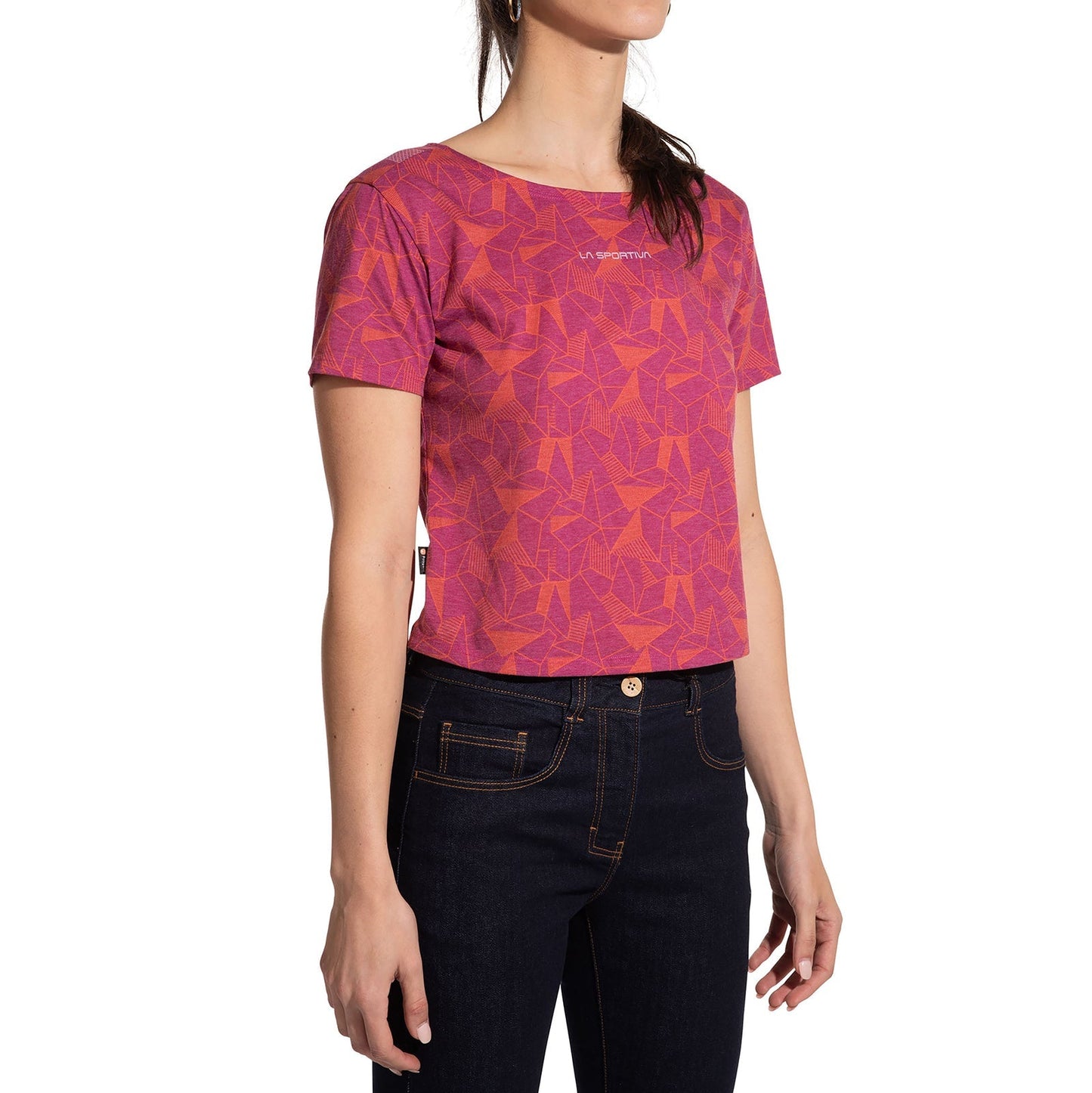 A woman wears the La Sportiva Dimension T-Shirt Women's (2025), a short-sleeved, pink and orange geometric crop top made from recycled polyester, with dark blue jeans against a plain white background.