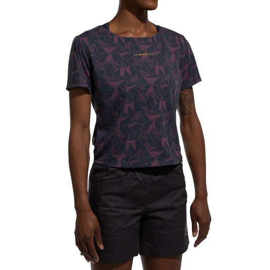 A person stands against a white background wearing the La Sportiva Dimension T-Shirt Women's (2025), a short-sleeve, patterned purple and navy top made from recycled polyester, and black shorts. The person's face is not visible.