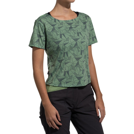 A woman stands against a white background wearing La Sportiva's Dimension T-Shirt Women's (2025), a green, geometric-patterned, short-sleeve top made from recycled polyester, paired with loose-fitting black pants.