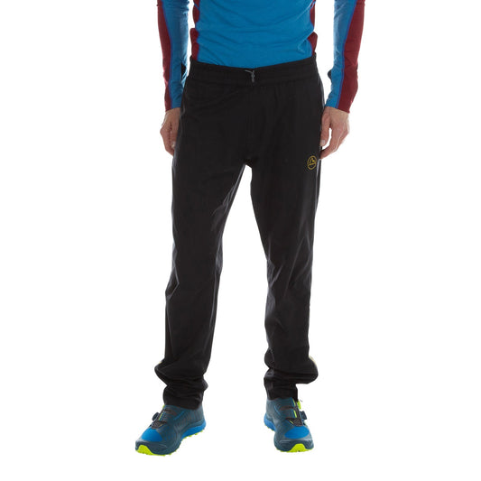 A person wearing a blue and maroon long-sleeve shirt, La Sportiva Drizzle Overpants Men's with a small yellow logo, and blue athletic shoes with yellow soles stands against a white background. The person's head is not visible.