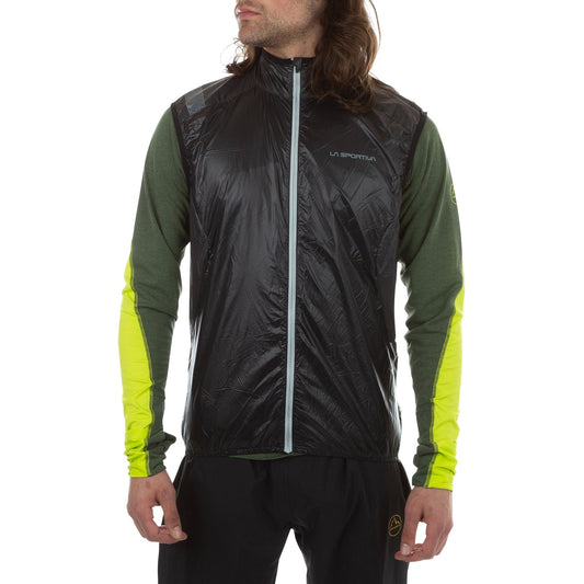 A man wears the La Sportiva Blizzard Windbreaker Vest in black over a green and yellow long-sleeve shirt with black pants, posing against a plain white background.
