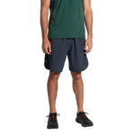 A person is wearing a green short-sleeve shirt, La Sportiva Sudden Shorts Men's navy running shorts with an 8" inseam, and black athletic shoes, standing against a white background. The head is not visible.