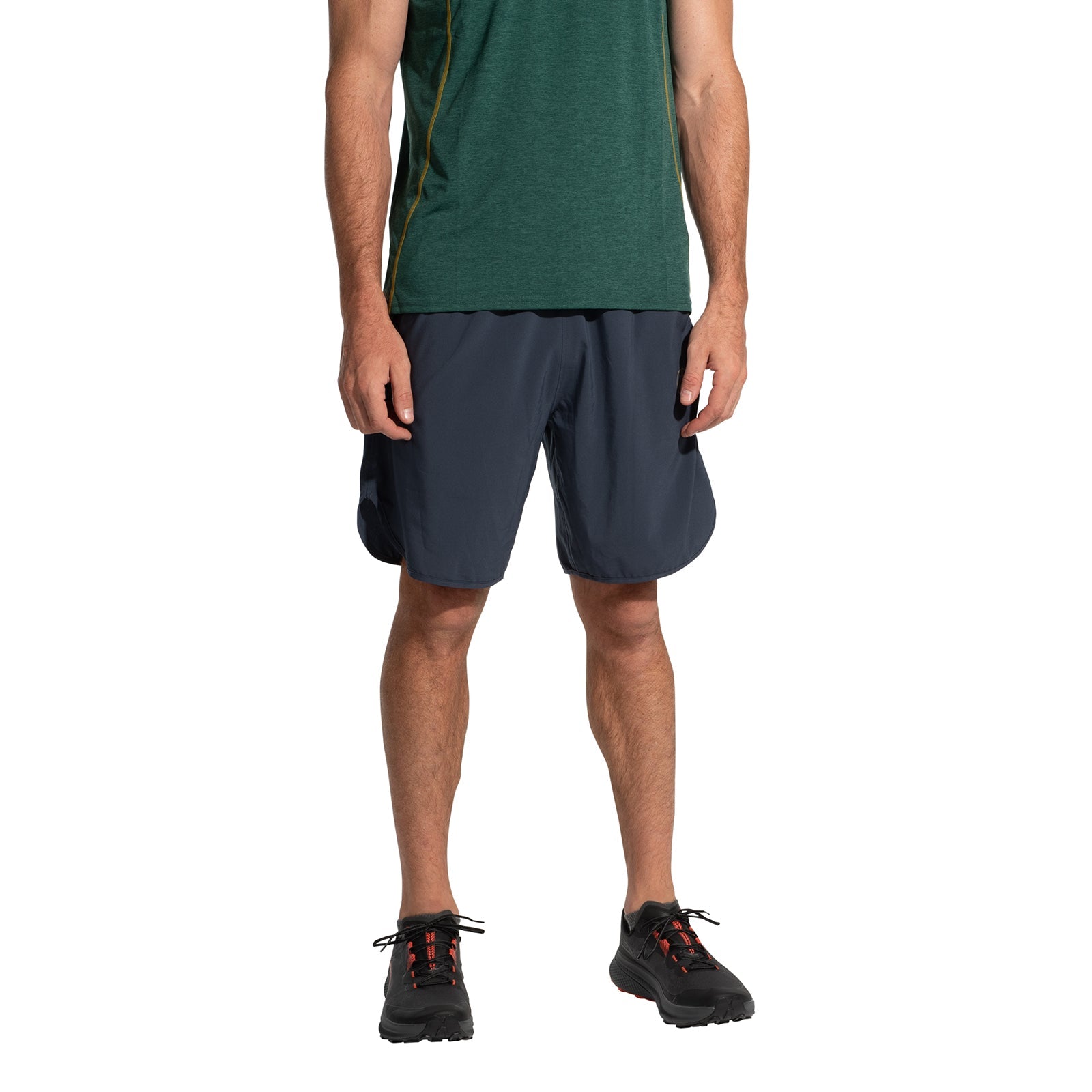 A person is wearing a green short-sleeve shirt, La Sportiva Sudden Shorts Men's navy running shorts with an 8" inseam, and black athletic shoes, standing against a white background. The head is not visible.