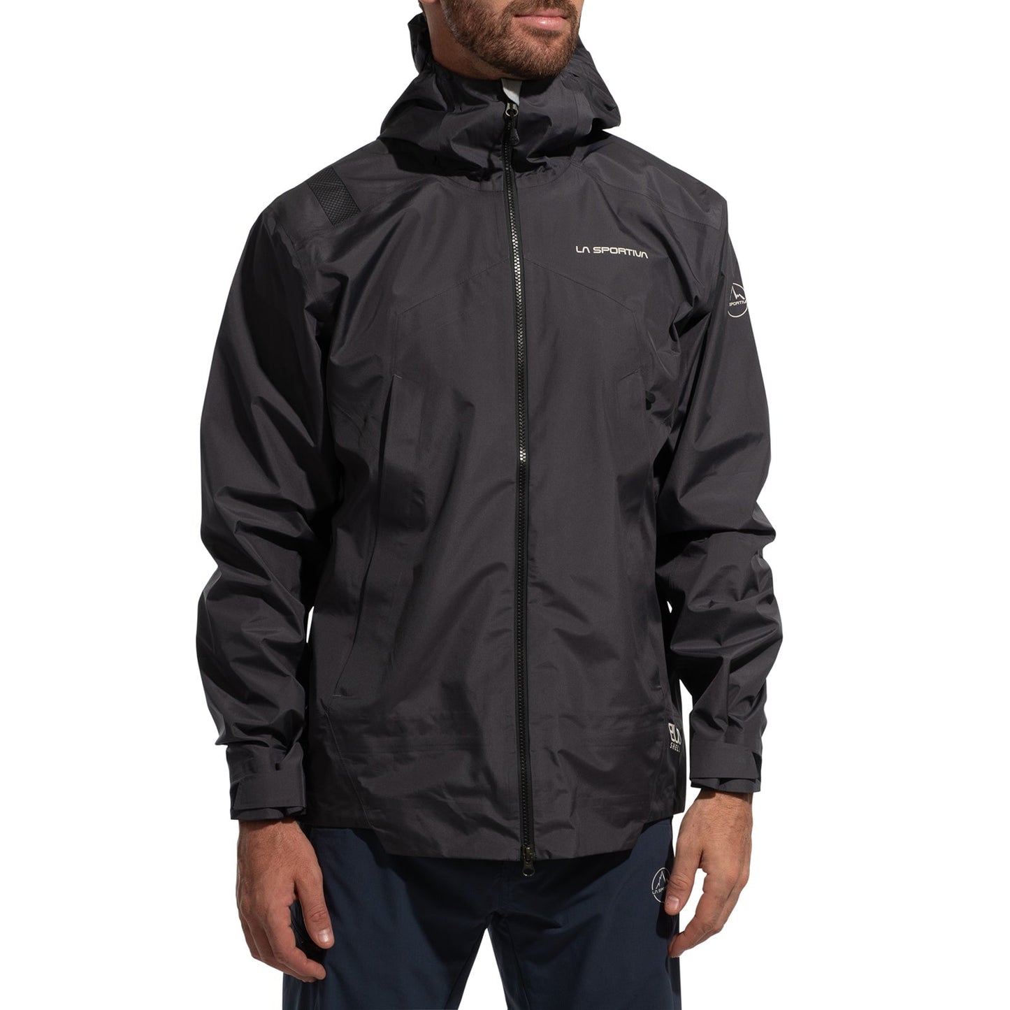 A man wears a black La Sportiva Discover Shell Jacket Men's (2025) with an adjustable hood, zippered front, and chest logo, standing against a white background.