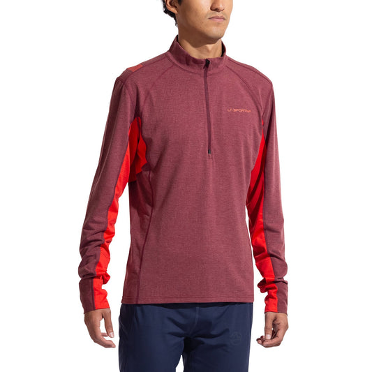 A man wearing the La Sportiva Swift Long Sleeve Men's shirt in maroon with red side panels and navy blue pants stands facing forward, arms relaxed—ideal attire for winter mountain running.