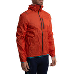 A man wearing the La Sportiva Ethereal Pro Jacket Men's—bright orange, hooded, with a front zipper and chest pocket, made from a lightweight three-layer shell—pairs it with dark athletic pants while standing against a white background.