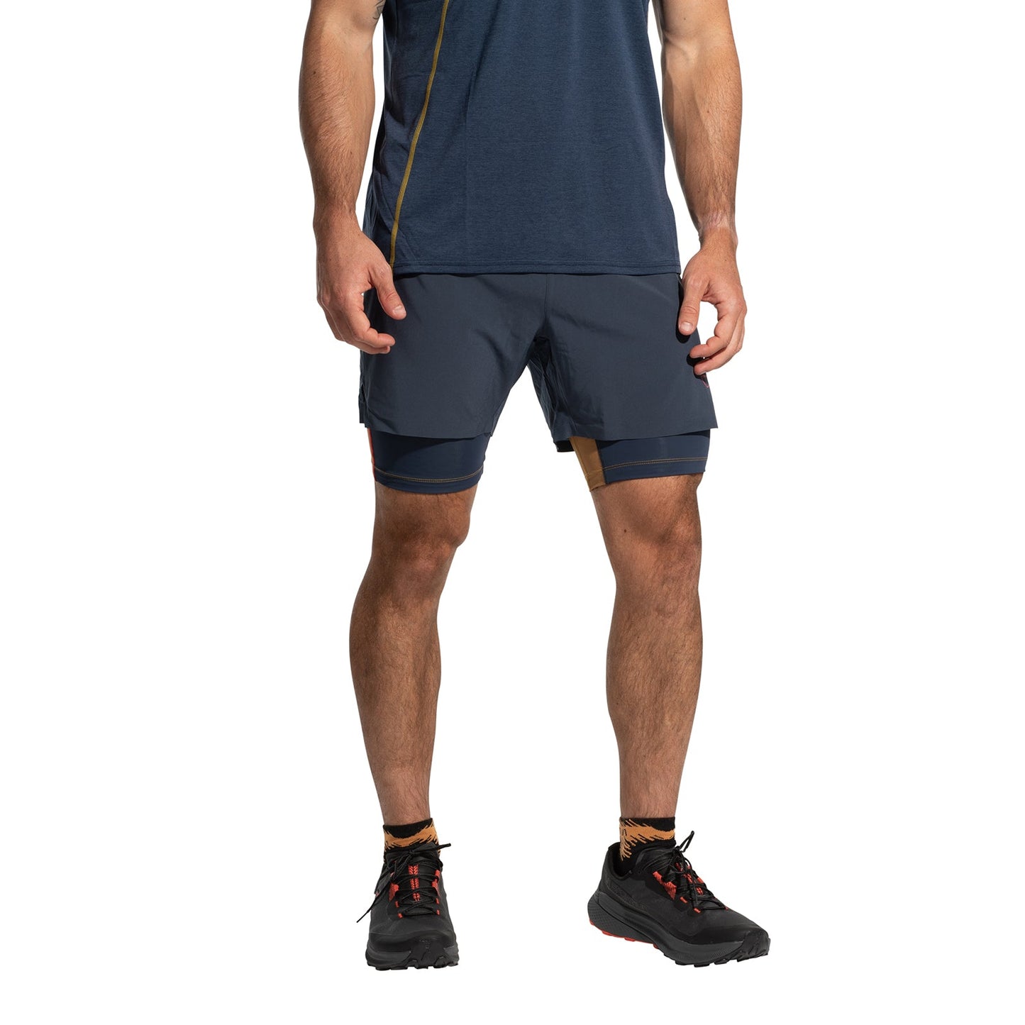 A man wears a navy athletic shirt and La Sportiva Ultra Distance Shorts Men's (2025) with built-in compression, paired with black trail running shoes. He faces forward, arms at his sides, head out of frame.