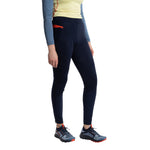 A woman wearing a light yellow long-sleeve top, La Sportiva Primal Pants Women's in dark navy with red accents for winter running, and gray trail running shoes with orange details, stands against a white background.