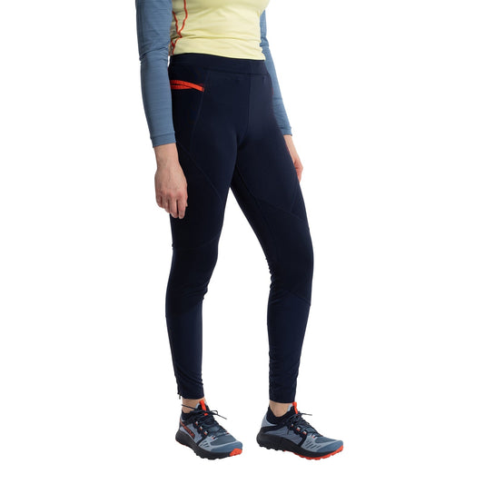 A woman wearing a light yellow long-sleeve top, La Sportiva Primal Pants Women's in dark navy with red accents for winter running, and gray trail running shoes with orange details, stands against a white background.
