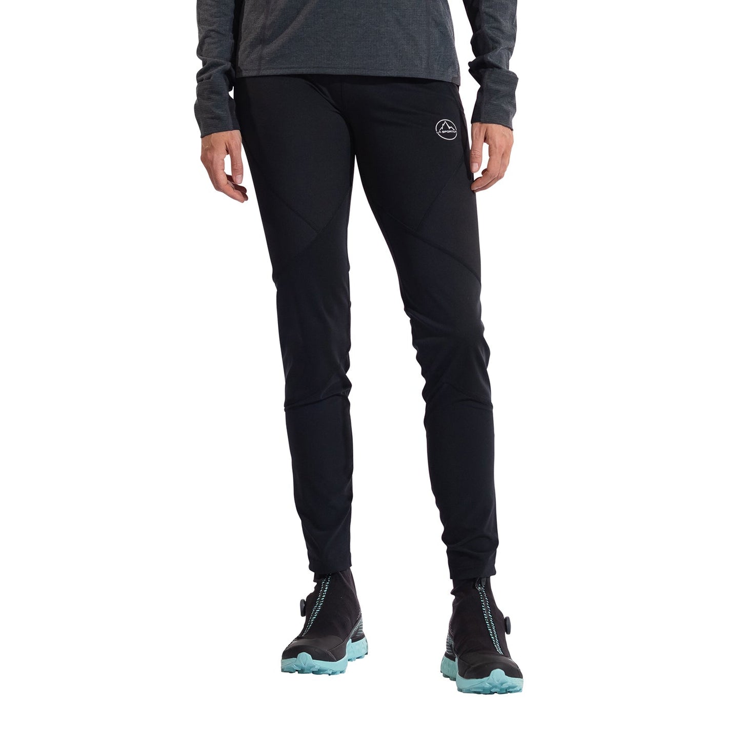 A woman wears La Sportiva Primal Pants Women's—black with a small white logo on the upper left thigh—paired with a gray long-sleeve top and black athletic shoes with light blue soles, ideal for winter runs against a white backdrop.