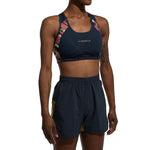 A person wearing the La Sportiva Hover Top Women's sports bra with colorful geometric straps and matching navy shorts with yellow side panels, standing against a white background.