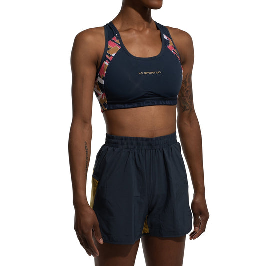 A person wearing the La Sportiva Hover Top Women's sports bra with colorful geometric straps and matching navy shorts with yellow side panels, standing against a white background.