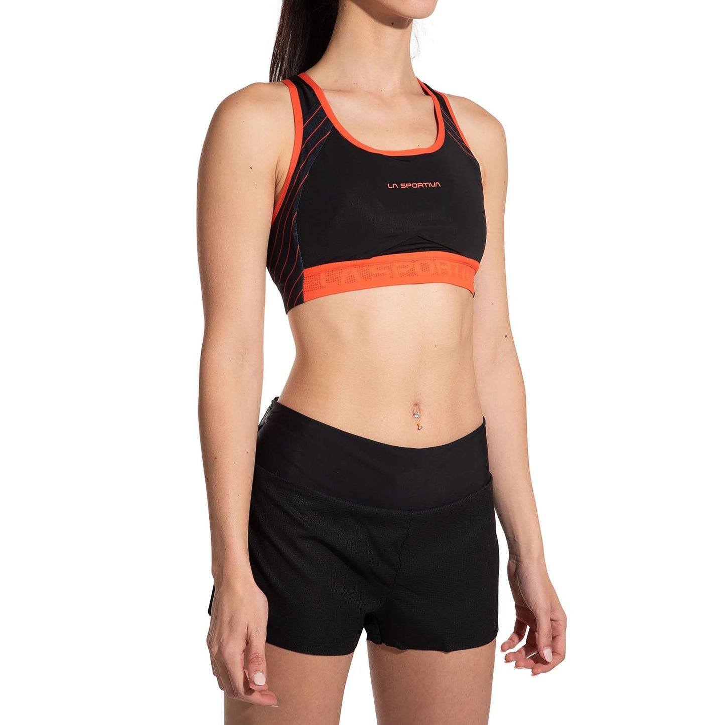 A woman models the La Sportiva Hover Top Women's sports bra in black and orange, paired with black athletic shorts, standing against a white background with her arms relaxed at her sides.