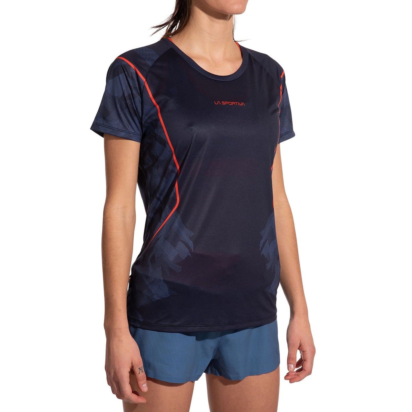 A woman wears the La Sportiva Pacer T-Shirt Women's—an ultralight running top crafted from recycled polyester with red accents—and blue shorts, standing with arms relaxed by her sides against a white background.