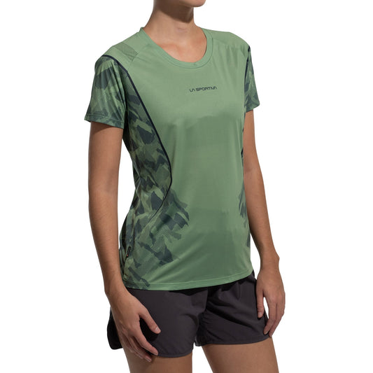 A woman wearing the La Sportiva Pacer T-Shirt Women's in green camo, made from recycled polyester, pairs it with black athletic shorts while standing against a plain white background.