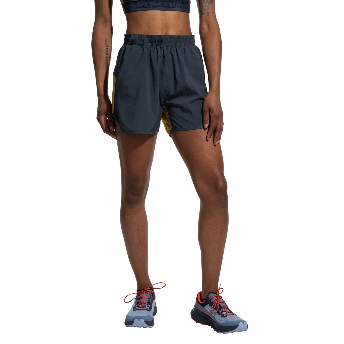 A person stands against a white background wearing La Sportiva Sudden Shorts Women's, featuring superlight fabric and an elastic waistband for comfort, paired with a sports bra and trail running shoes with red laces. Face not visible.