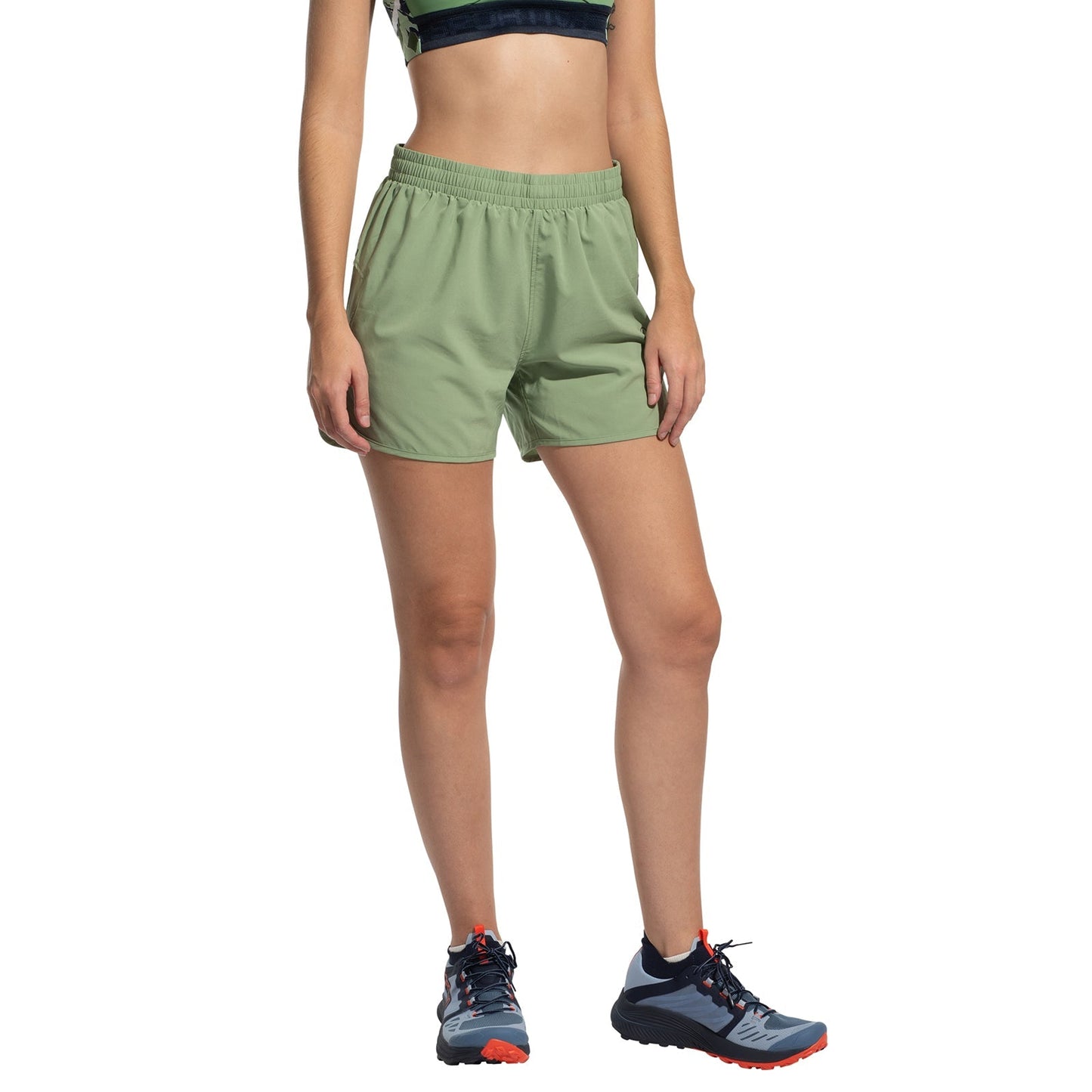 A woman wearing La Sportiva Sudden Shorts in light green stands against a white background, paired with a dark sports bra and blue trail running shoes with orange laces.