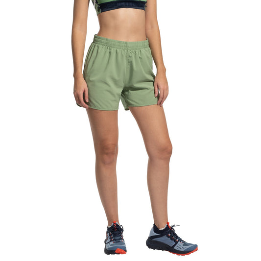 A woman wearing La Sportiva Sudden Shorts in light green stands against a white background, paired with a dark sports bra and blue trail running shoes with orange laces.