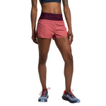 A person stands facing forward, wearing La Sportiva Freccia Shorts Women's in pink with a maroon waistband. Their navy sports bra and blue trail running shoes with red laces are also visible. Only their body from the shoulders down is shown.