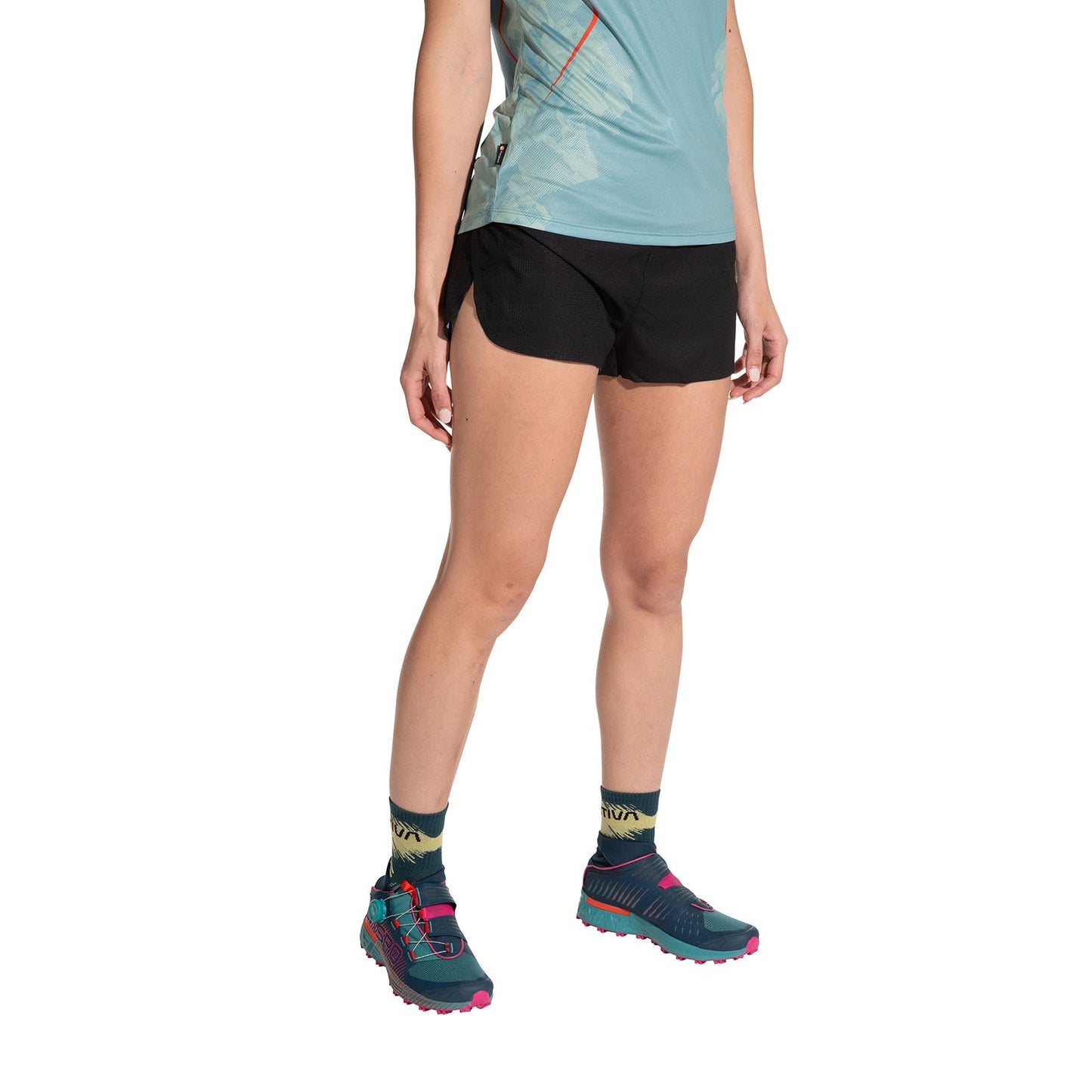 Against a white background, a person wears a light blue athletic shirt, La Sportiva Freccia Shorts Women's in black, patterned socks, and teal trail running shoes with pink accents. The person's face is not visible.