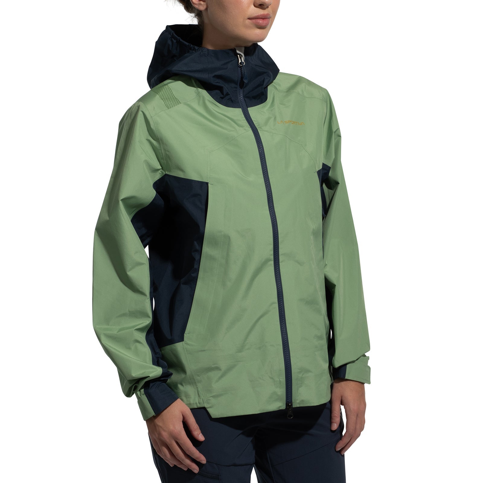 A woman wears the La Sportiva Discover Shell Jacket (2025) featuring a light green front and sleeves, dark blue side panels, adjustable hood, and front zipper, standing against a plain white backdrop.