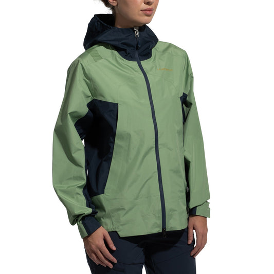 A woman wears the La Sportiva Discover Shell Jacket (2025) featuring a light green front and sleeves, dark blue side panels, adjustable hood, and front zipper, standing against a plain white backdrop.