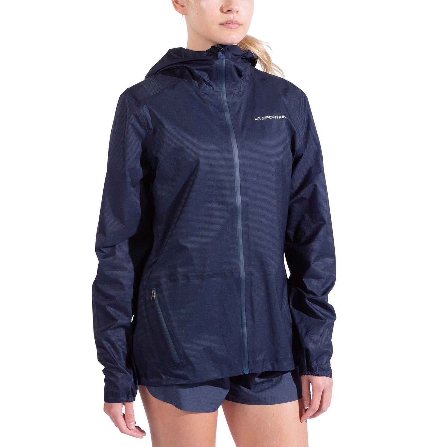 A woman stands against a plain white background wearing the La Sportiva Ethereal Pro Jacket Women's in navy blue, featuring a lightweight three-layer shell, front zipper, zippered pockets, and a small chest logo.