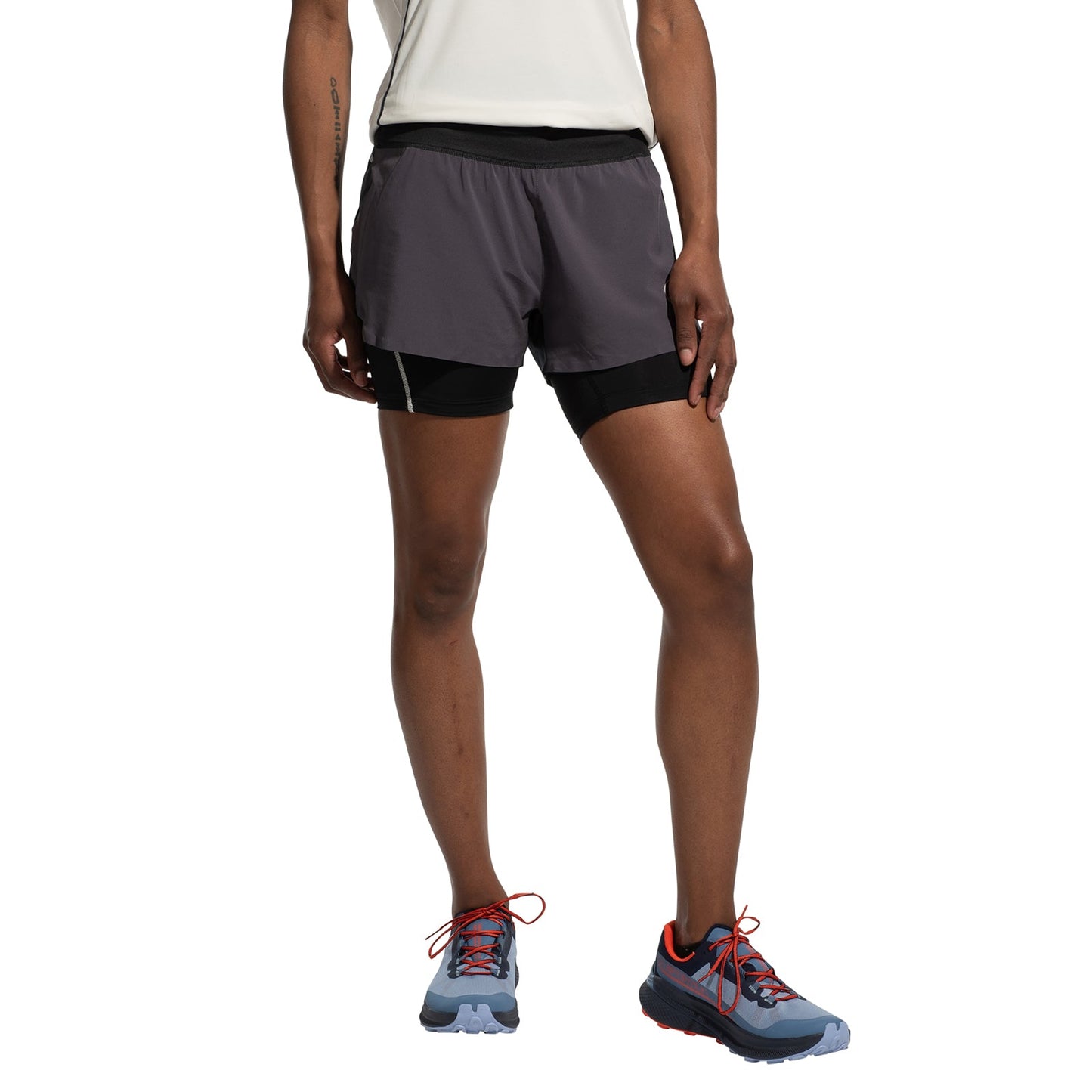 A woman wearing a white shirt, La Sportiva Ultra Distance Shorts Women's with black inner shorts, and blue trail running shoes with orange laces stands against a white background.