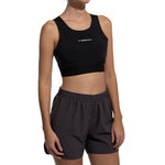 A woman wearing the La Sportiva Captive Air Top Women's (2025), a black, medium-support sports bra, and dark gray recycled-fabric athletic shorts stands against a plain white background with her face partially out of frame.
