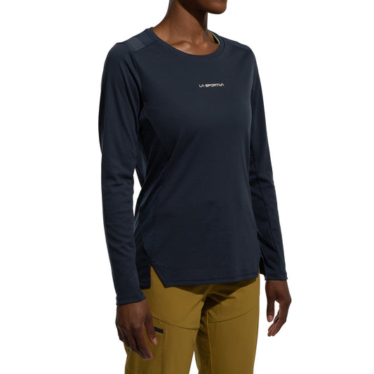 A woman wears the La Sportiva Beyond Long Sleeve Women's (2025) in dark blue Polartec Delta Cooling Fabric and mustard yellow pants, standing against a plain white background.