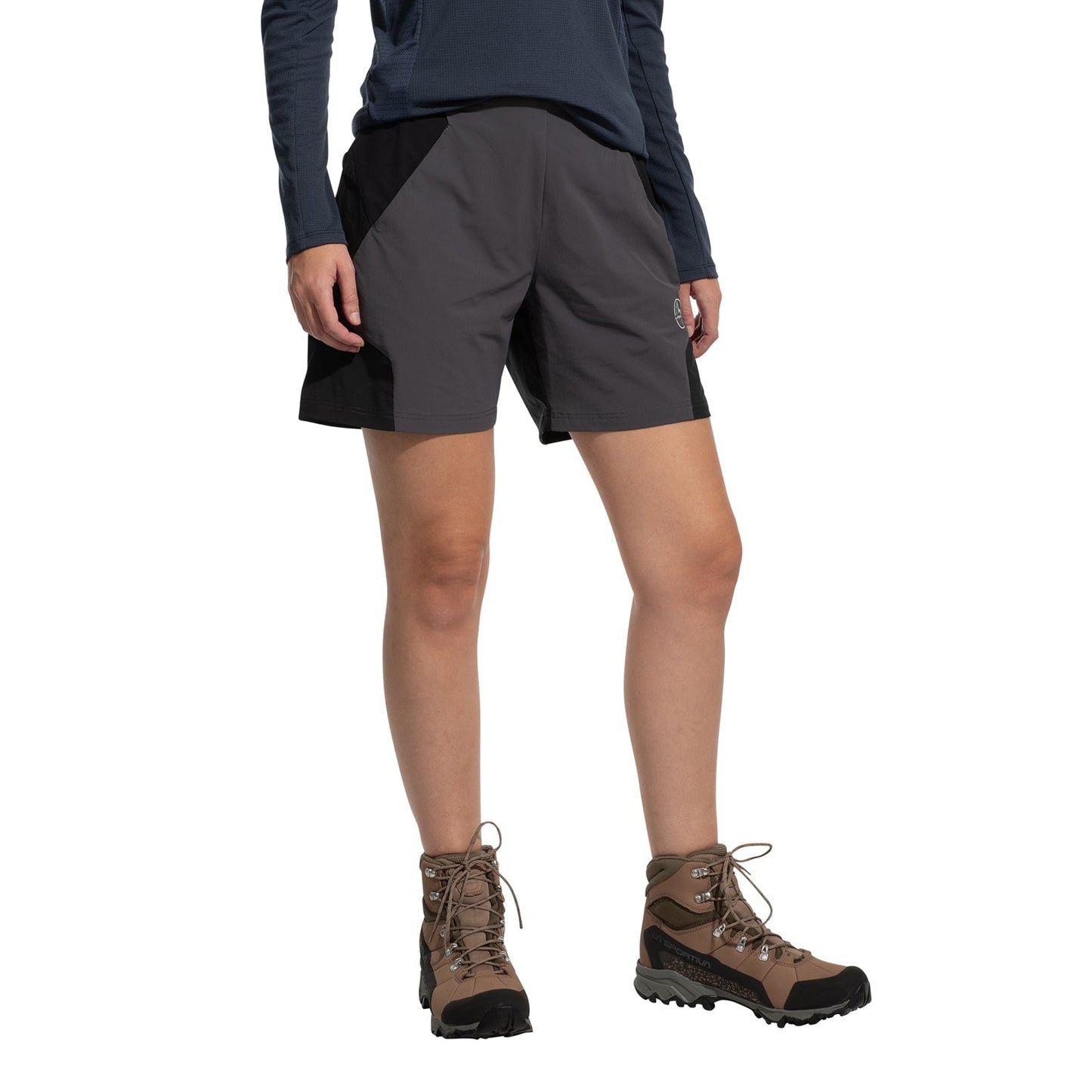 A person wears La Sportiva Trail Guard Shorts Women's (2025), paired with a navy long-sleeve shirt and brown hiking boots, standing against a white background. The image highlights the shorts, perfect for fast-hiking adventures.