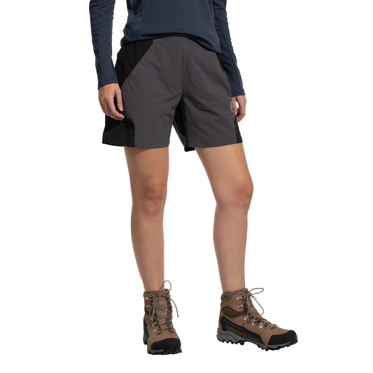 A person wears La Sportiva Trail Guard Shorts Women's (2025), paired with a navy long-sleeve shirt and brown hiking boots, standing against a white background. The image highlights the shorts, perfect for fast-hiking adventures.