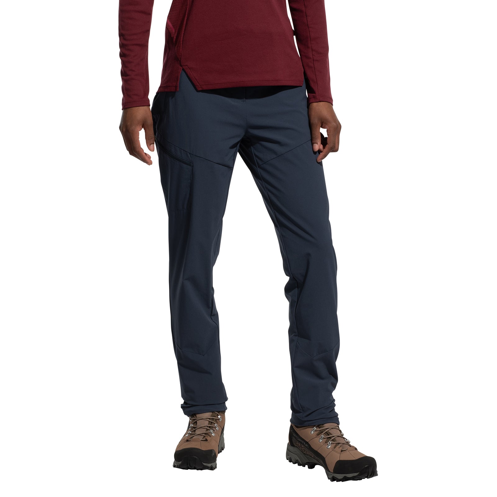 A person is wearing La Sportiva's West Crest Pants Women's (2025), which are dark blue hiking pants with zippered pockets. They also have on a burgundy long-sleeve shirt and brown hiking boots, standing against a white background, head not visible.