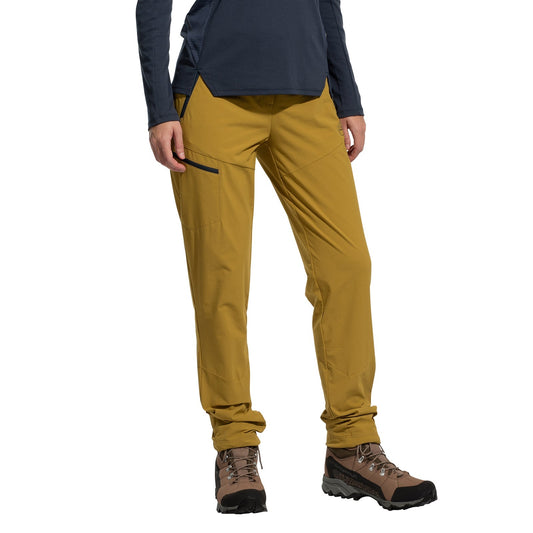 A person wears La Sportiva West Crest Pants Women's (2025) in mustard yellow, featuring zippered pockets, paired with a navy long-sleeve top and brown hiking boots, standing against a plain white background.