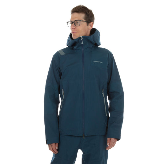 A man in glasses wears a blue La Sportiva Roseg GTX Jacket Men's with a zipper and matching pants, facing slightly to the side against a plain white background.