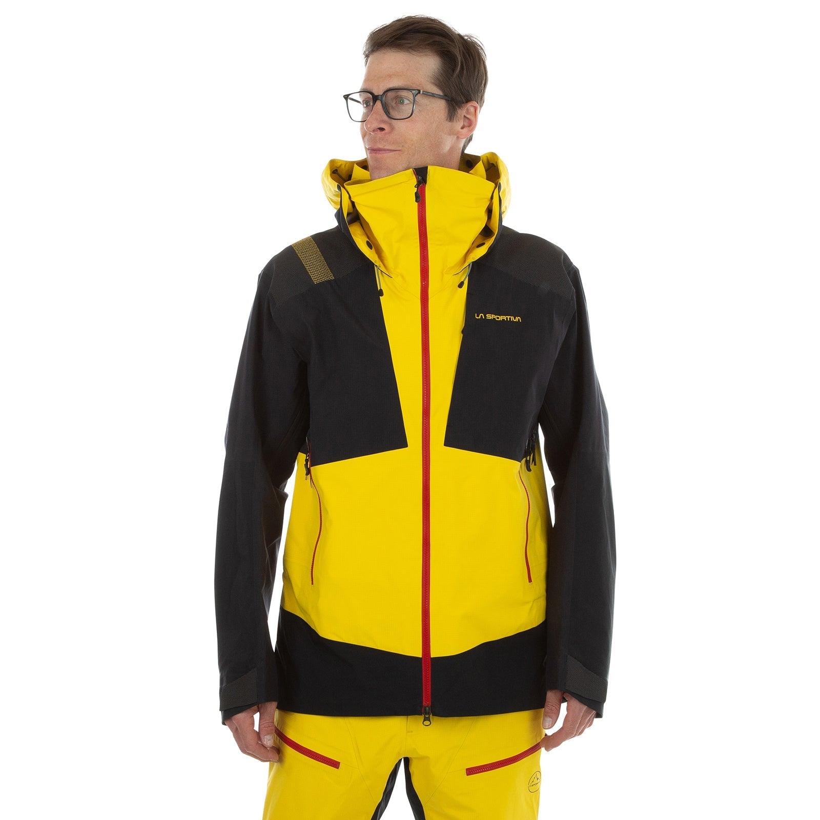 A man wearing glasses models the La Sportiva Supercouloir GTX Pro Jacket Men's (2024) in yellow and black with a hood, red zipper, and La Sportiva logo on the chest. He stands against a white background—ideal for ice climbing or as a winter shell.