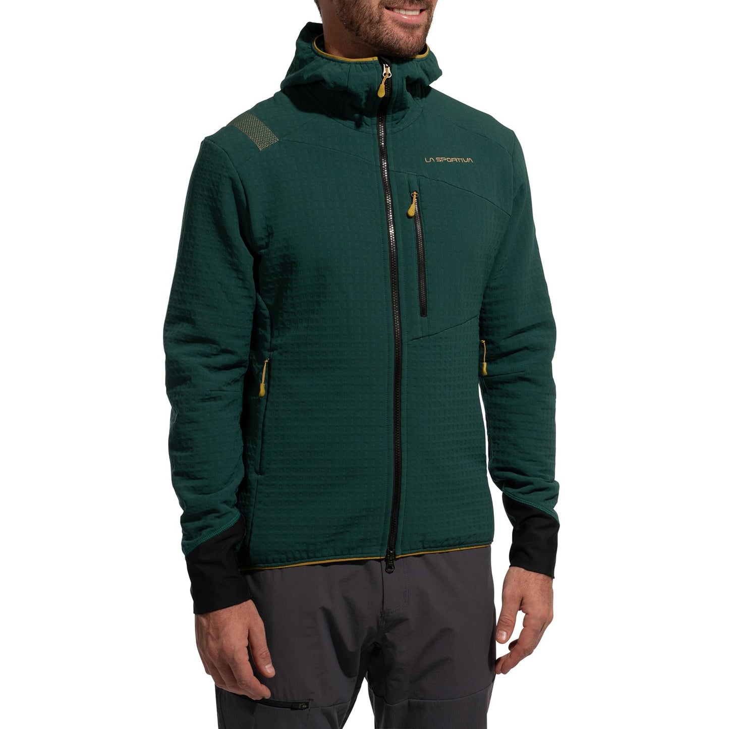 A man wearing the La Sportiva Bristen Thermal Hoody Men's in dark green, with yellow zipper accents and a small chest logo, smiles while facing slightly to the side. Designed for outdoor activities, he pairs it with black cuffs and gray pants.