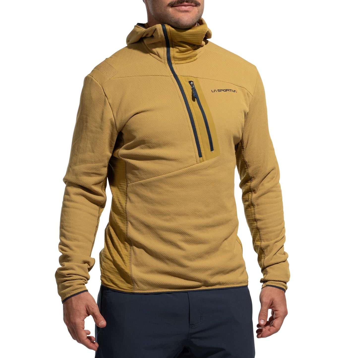 A person wears the La Sportiva Big Wall Pullover Hoody Men's (2025), a tan climbing hoody with Polartec inserts and a zippered chest pocket with logo, paired with dark blue pants, standing against a plain white background.