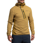 A person wears the La Sportiva Big Wall Pullover Hoody Men's (2025), a tan climbing hoody with Polartec inserts and a zippered chest pocket with logo, paired with dark blue pants, standing against a plain white background.