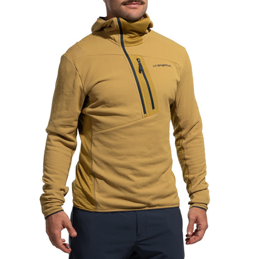 A person wears the La Sportiva Big Wall Pullover Hoody Men's (2025), a tan climbing hoody with Polartec inserts and a zippered chest pocket with logo, paired with dark blue pants, standing against a plain white background.