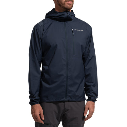 A person stands with arms relaxed, wearing the La Sportiva Wall Breeze Stretch Jacket Men's (2025) in dark navy blue. The alpine jacket features a front zipper, chest pocket, helmet-compatible hood, wind protection, and a small chest logo.
