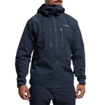 A person wears a dark blue La Sportiva Helixir Shell Jacket Men's (2025) with matching pants. The jacket has Spectra fibers, a front zipper, two chest zippered pockets, and a small chest logo. Only the lower half of their face is visible.