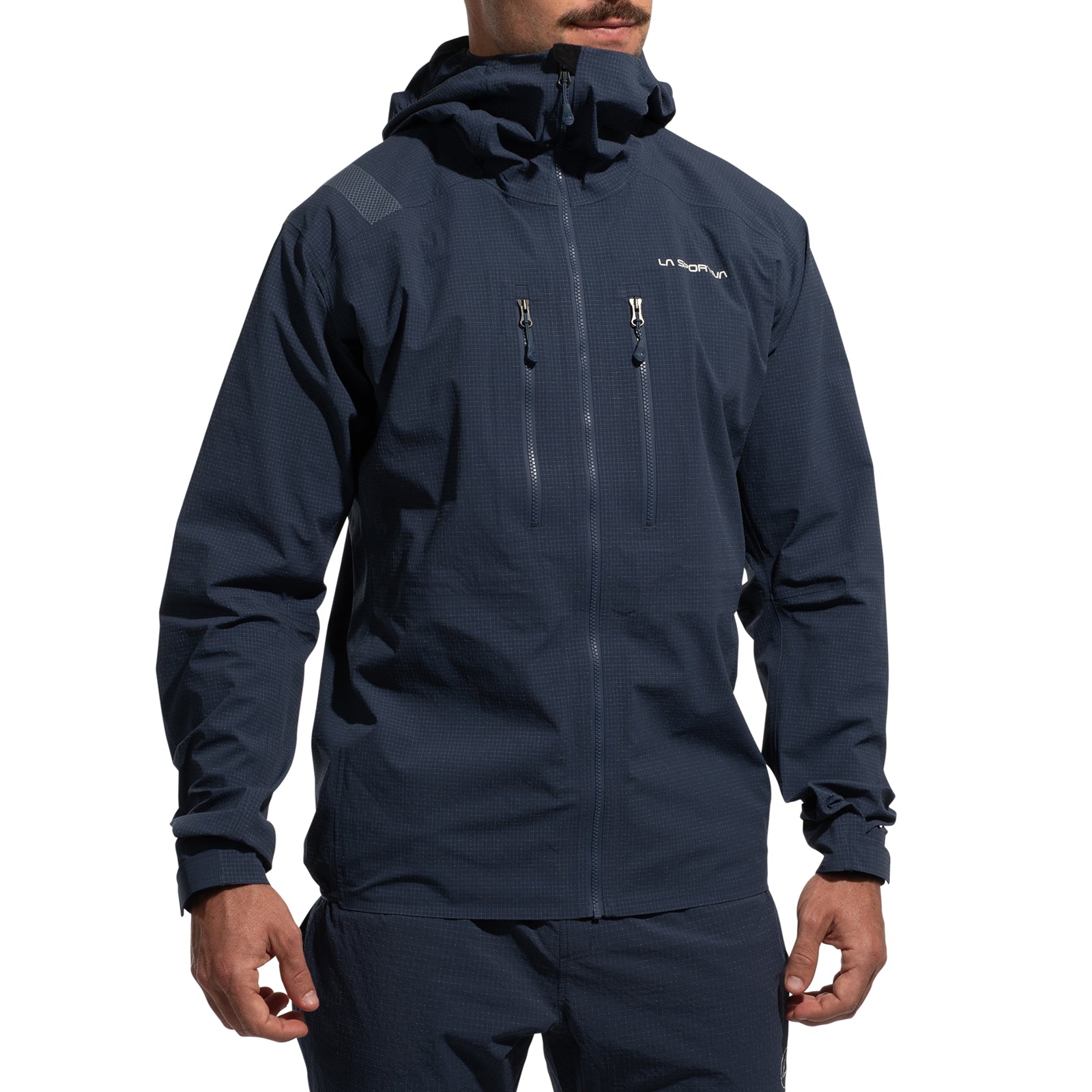 A person wears a dark blue La Sportiva Helixir Shell Jacket Men's (2025) with matching pants. The jacket has Spectra fibers, a front zipper, two chest zippered pockets, and a small chest logo. Only the lower half of their face is visible.