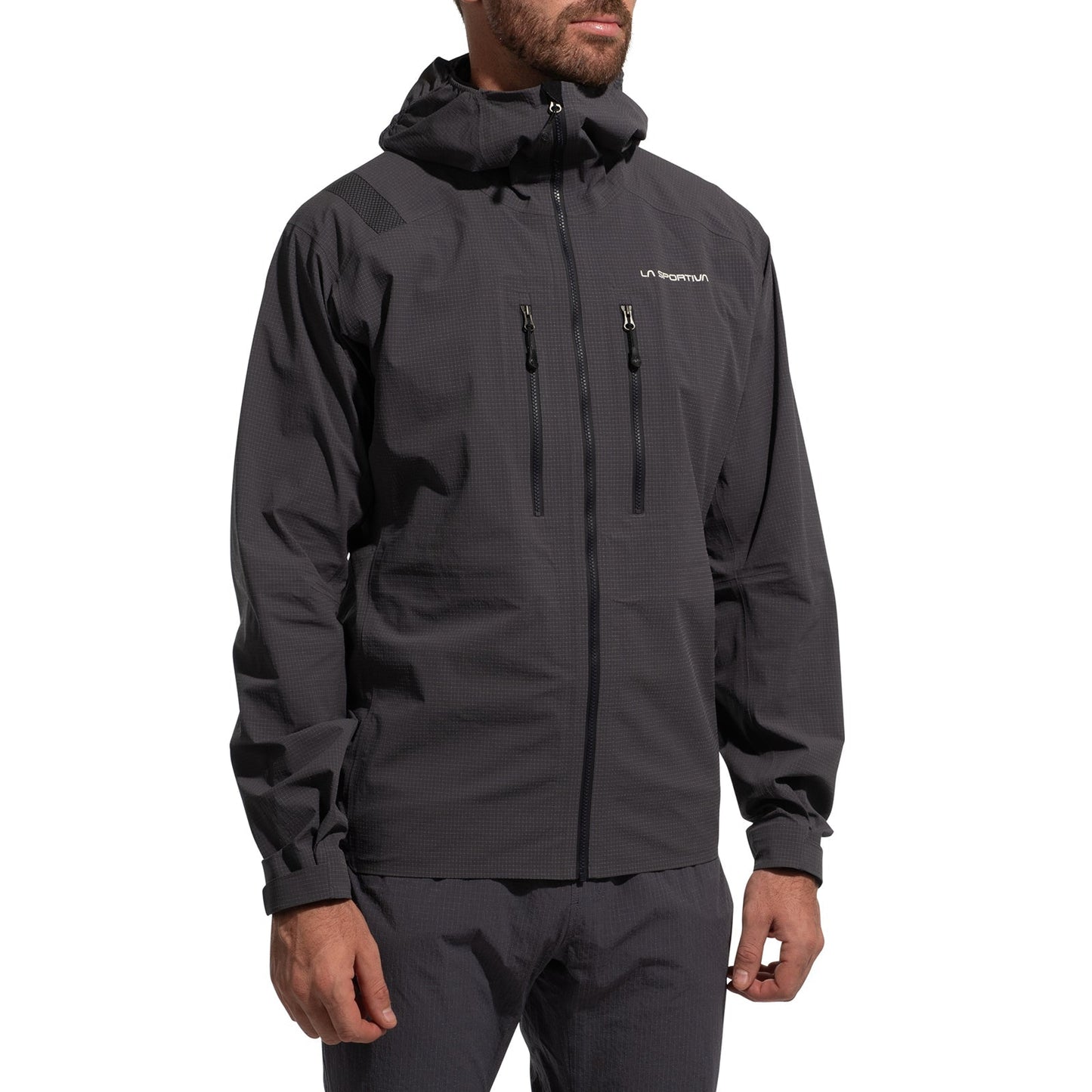 A man stands with arms relaxed, wearing the La Sportiva Helixir Shell Jacket Men's (2025) in dark gray with zippered chest pockets, matching pants, and a subtle logo on the left chest.