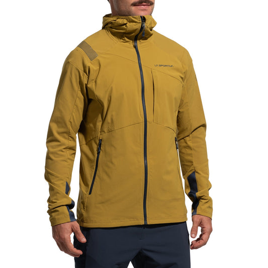 A man stands with hands at his sides, wearing the La Sportiva Aequilibrium Speed Jacket Men's (2025)—a mustard yellow, lightweight hooded jacket with a front zipper, dark blue accents, and branded chest detailing.